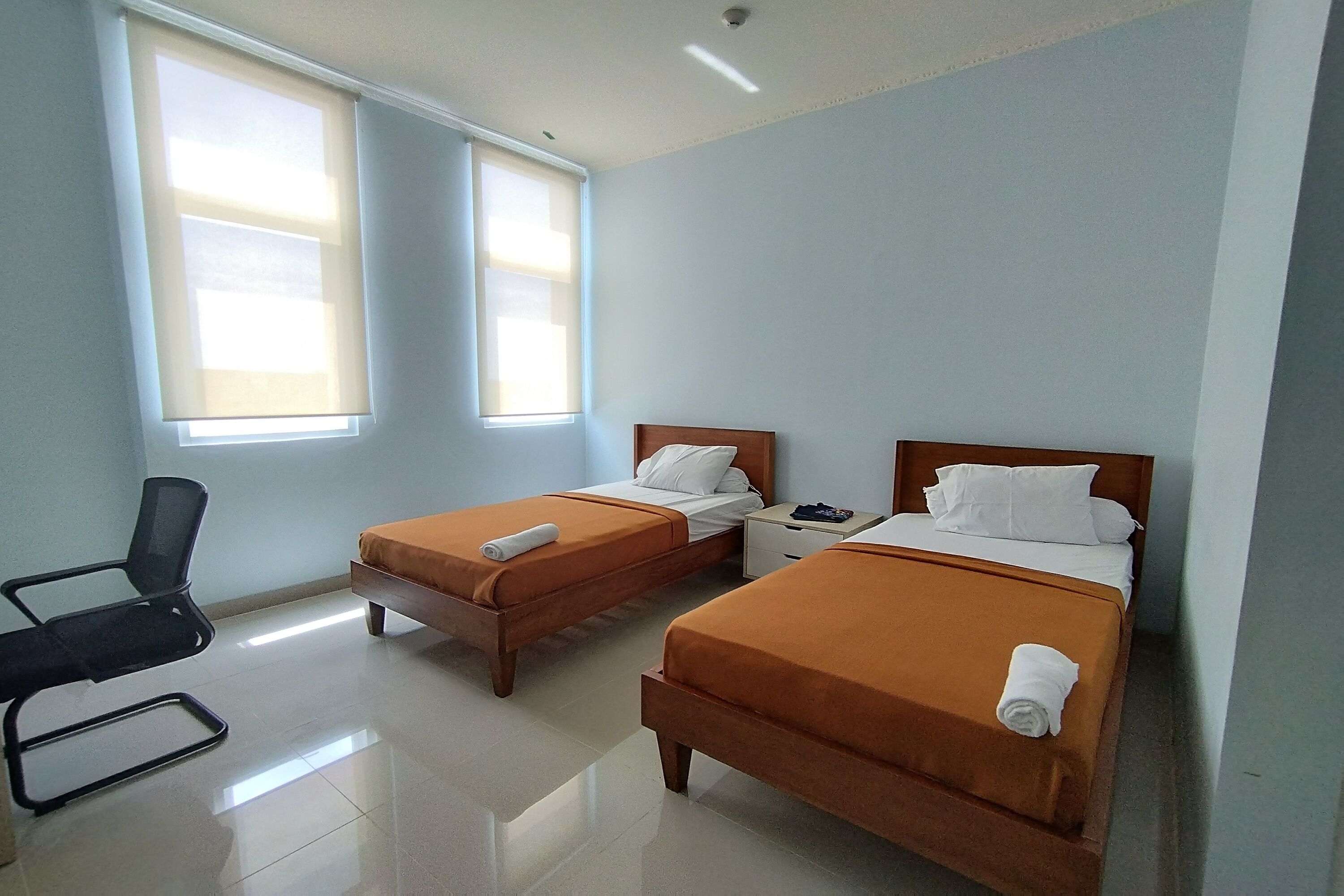 Une chambre d’hôtel avec deux lits simples, une chaise, une table de chevet et une fenêtre avec des stores.