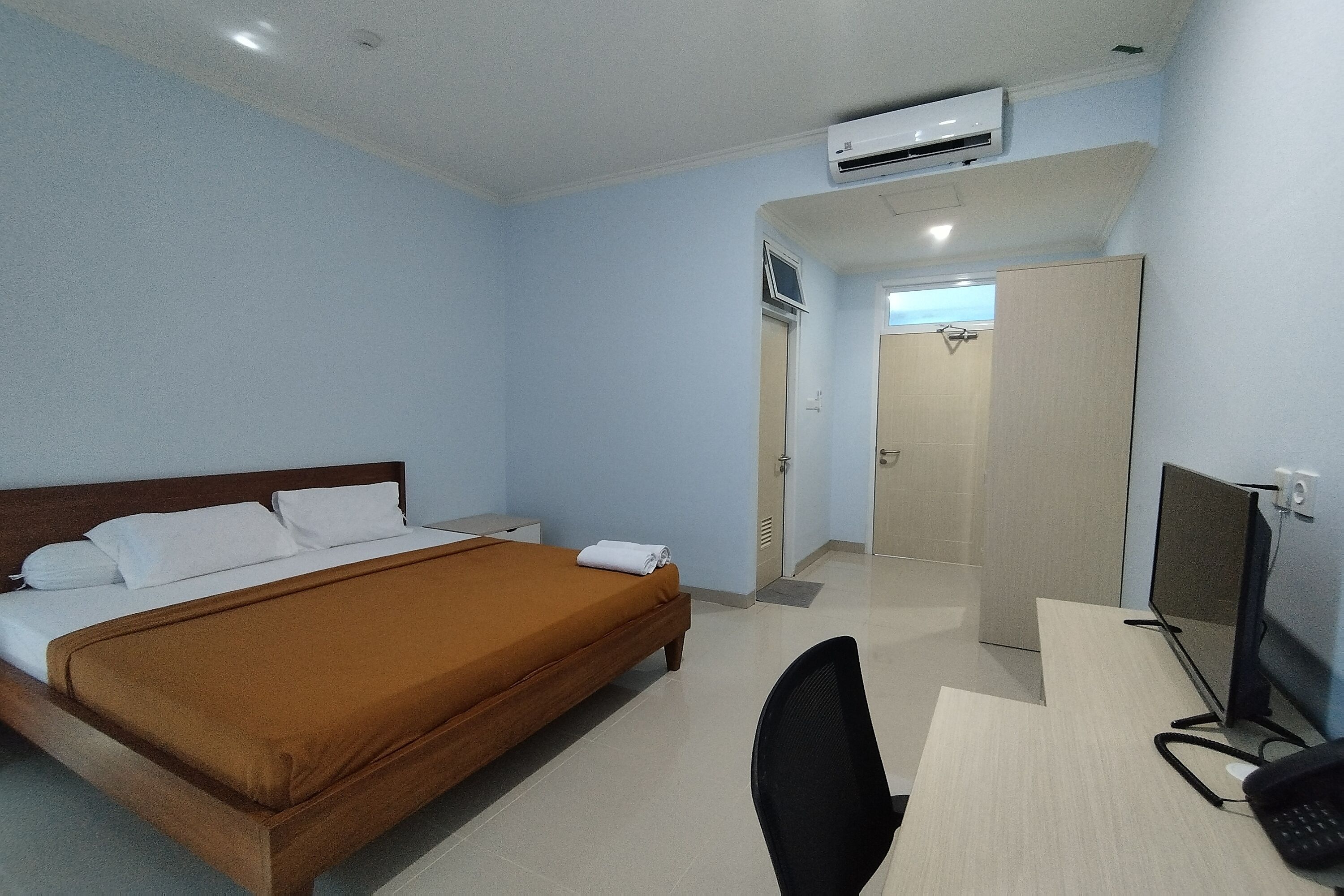 A hotel room with a bed, desk, chair, and an air conditioning unit.