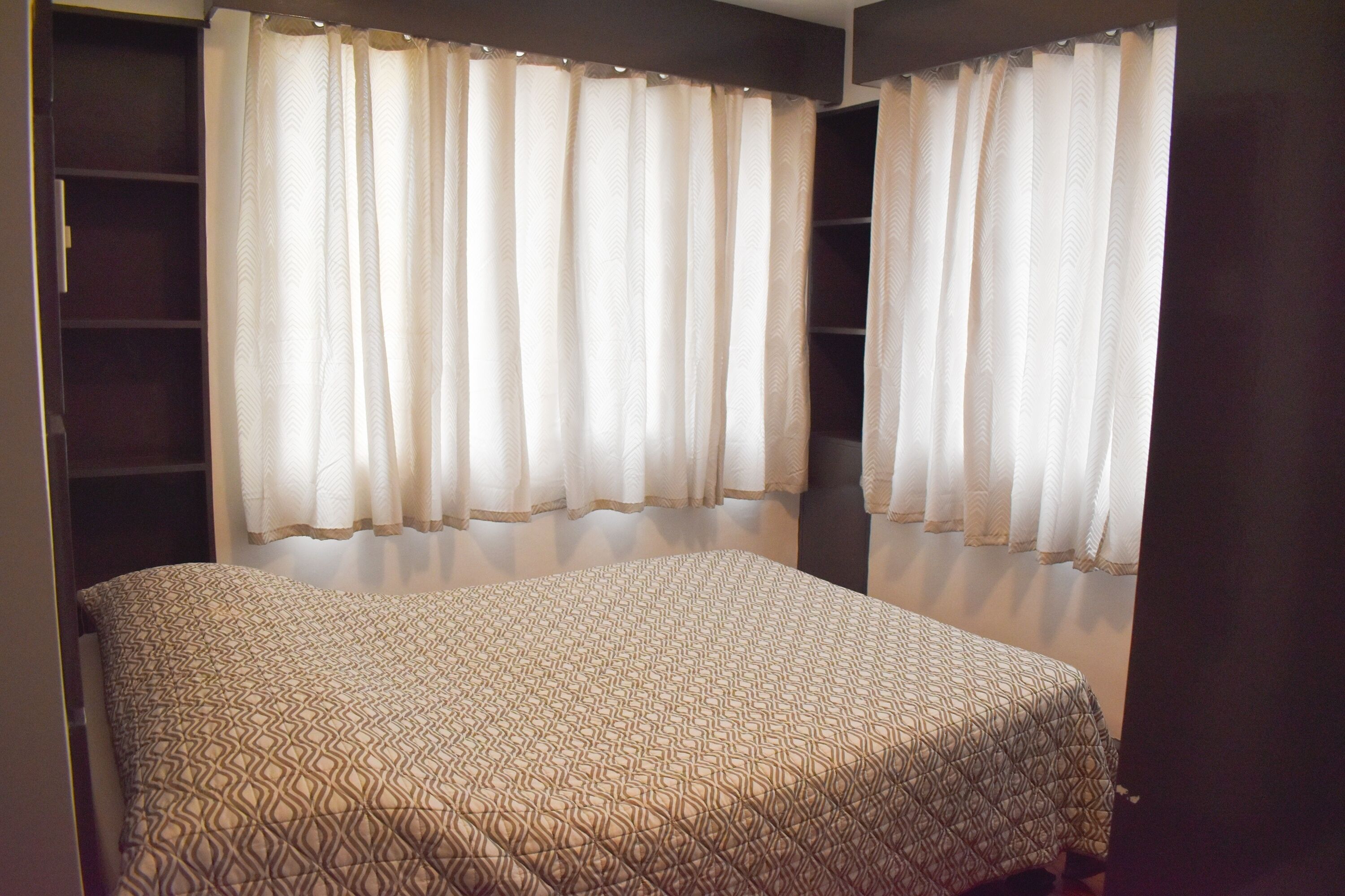 A bedroom with a bed, two windows with curtains, and a wardrobe.