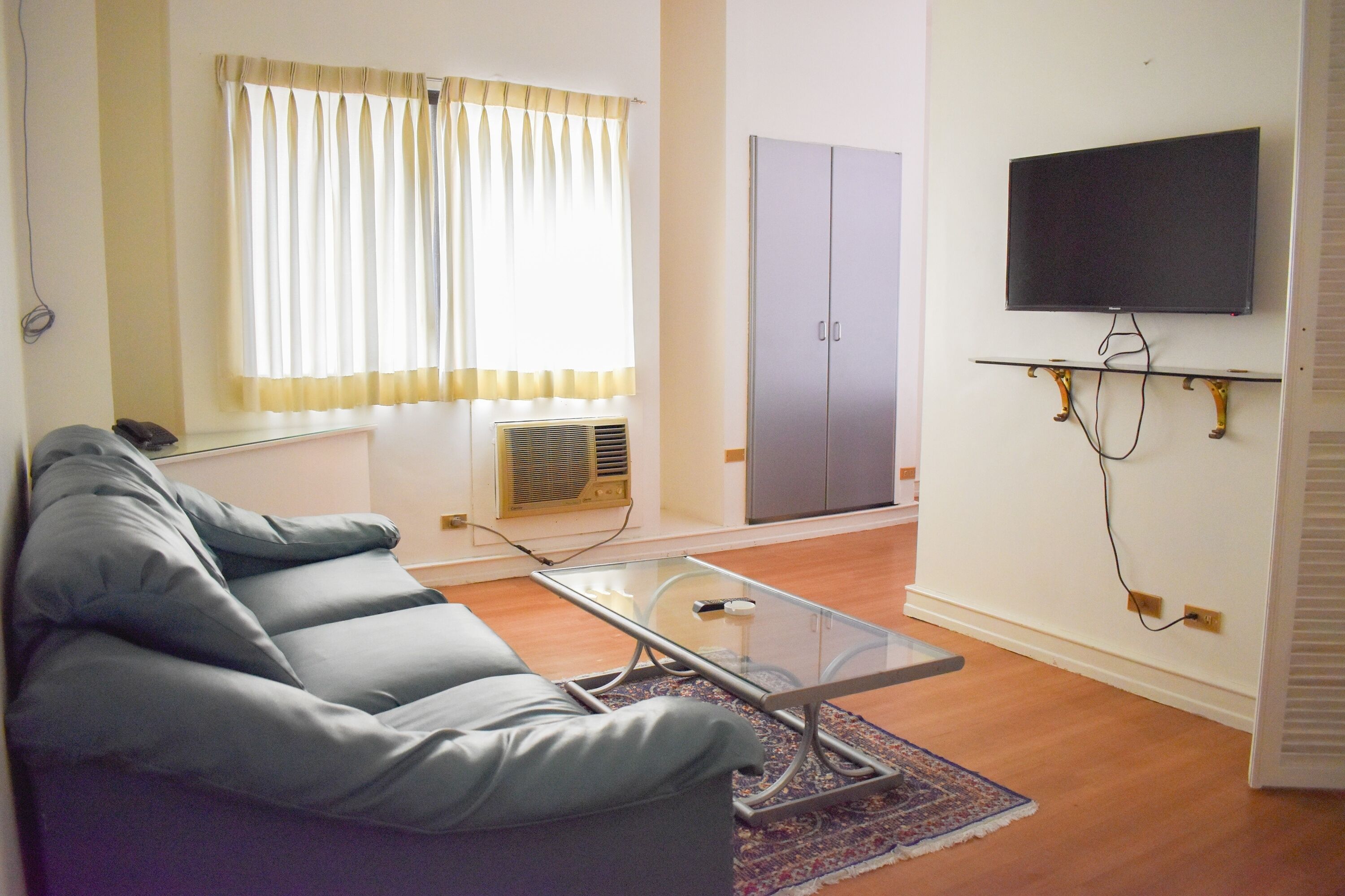 A living room with a grey sofa, a glass coffee table, a television mounted on the wall, a wardrobe, and a radiator.