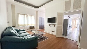 Un salon avec un canapé bleu, une table basse en verre, un téléviseur à écran plat fixé au mur et une cuisine visible à travers une porte ouverte.