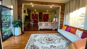 A spacious living room with a large rug, a sofa with colorful cushions, and a wooden coffee table. There is a dining area with a red cabinet and a kitchen in the background. - Baan Wang Tarn (Hang Dong)