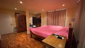 A hotel room with a wooden floor, a bed with a pink bedspread, a wooden nightstand, a television, and a wardrobe. - Baan Wang Tarn (Hang Dong)