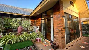A modern house with a brick exterior, large glass doors, and a well-maintained garden with various plants and a small pink stool. - Baan Wang Tarn (Hang Dong)