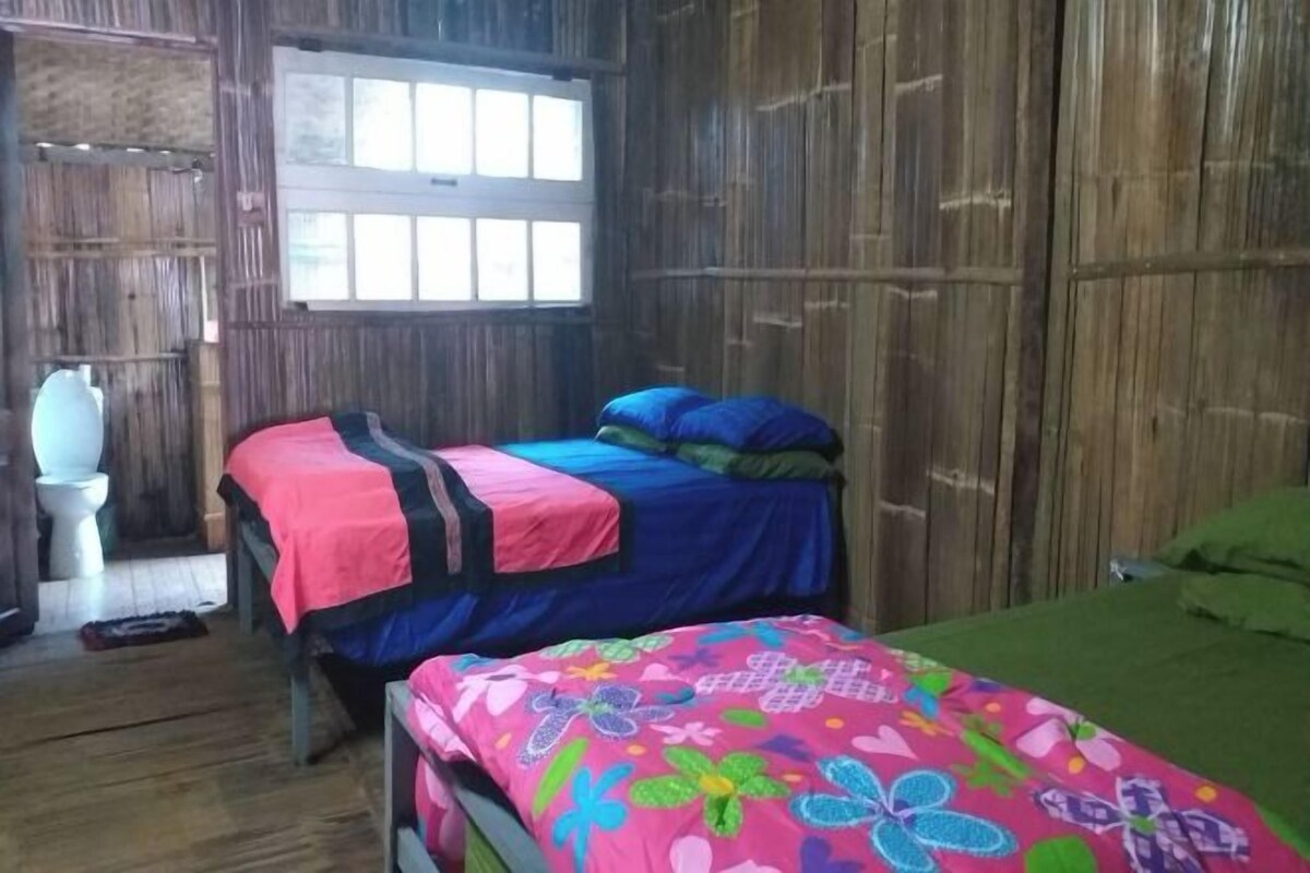 A room with two beds, one with a colorful floral bedspread and another with blue bedding. There is a window with blinds, a white toilet, and a wooden wall.