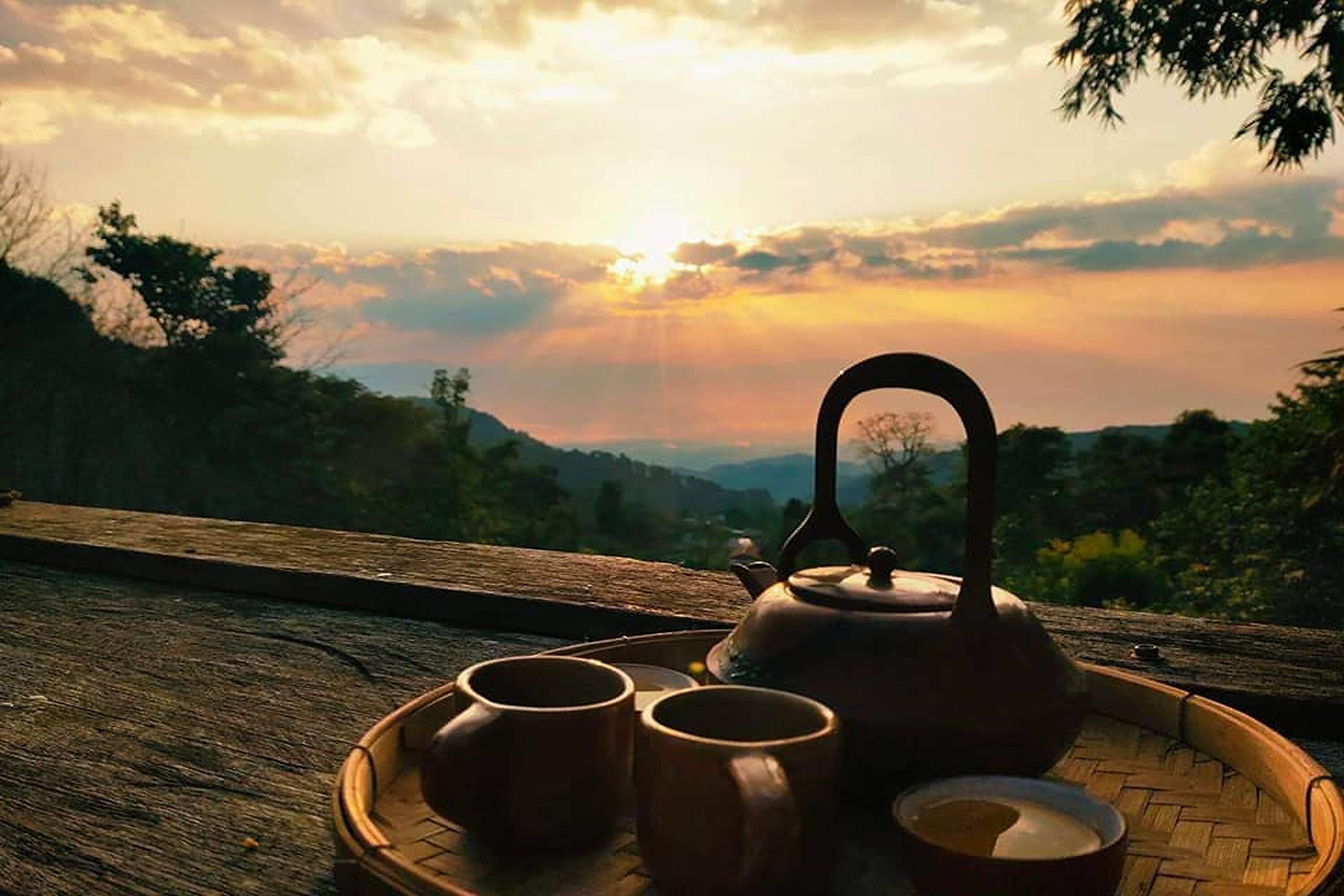 A wooden tray with a teapot and two cups on a wooden surface with a scenic view in the background.