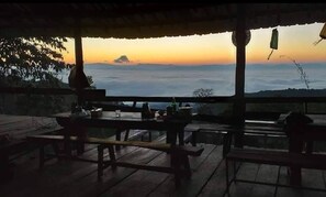An outdoor seating area with wooden tables and benches, overlooking a scenic view of a sunset over a body of water. - Doi Pumuen Lahu Homestay (Mae Chan)