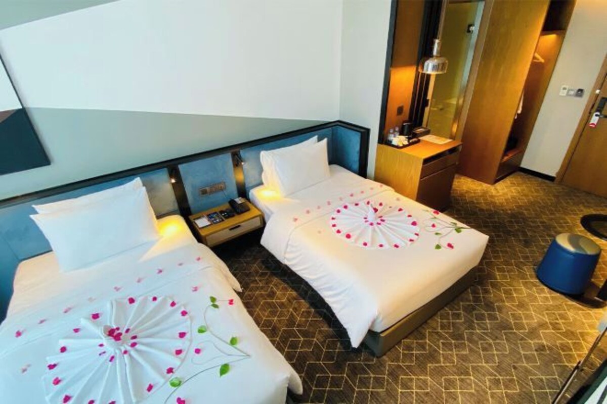 Une chambre d’hôtel avec deux lits, chacun recouvert d’une courtepointe blanche et agrémenté de coussins décoratifs. On y trouve une petite table de chevet avec une lampe, un miroir et une armoire en bois. Le sol de la chambre est orné d’un motif géométrique et un grand miroir est accroché au mur.
