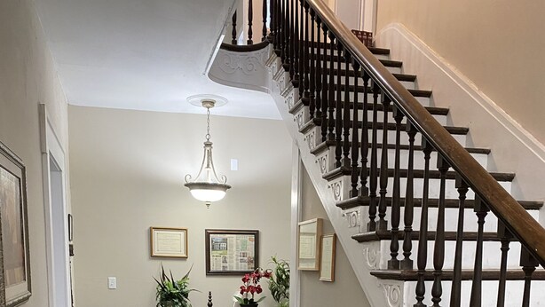 A staircase with a wooden handrail and balusters, leading to a landing with a chandelier, framed certificates, and a wooden bench with red cushions. - Historic McDonald Airbnb (Attica)