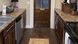 A kitchen with a wooden door, a clock, shelves with dishes and plants, a coffee maker, and a rug. - Historic McDonald Airbnb (Attica)
