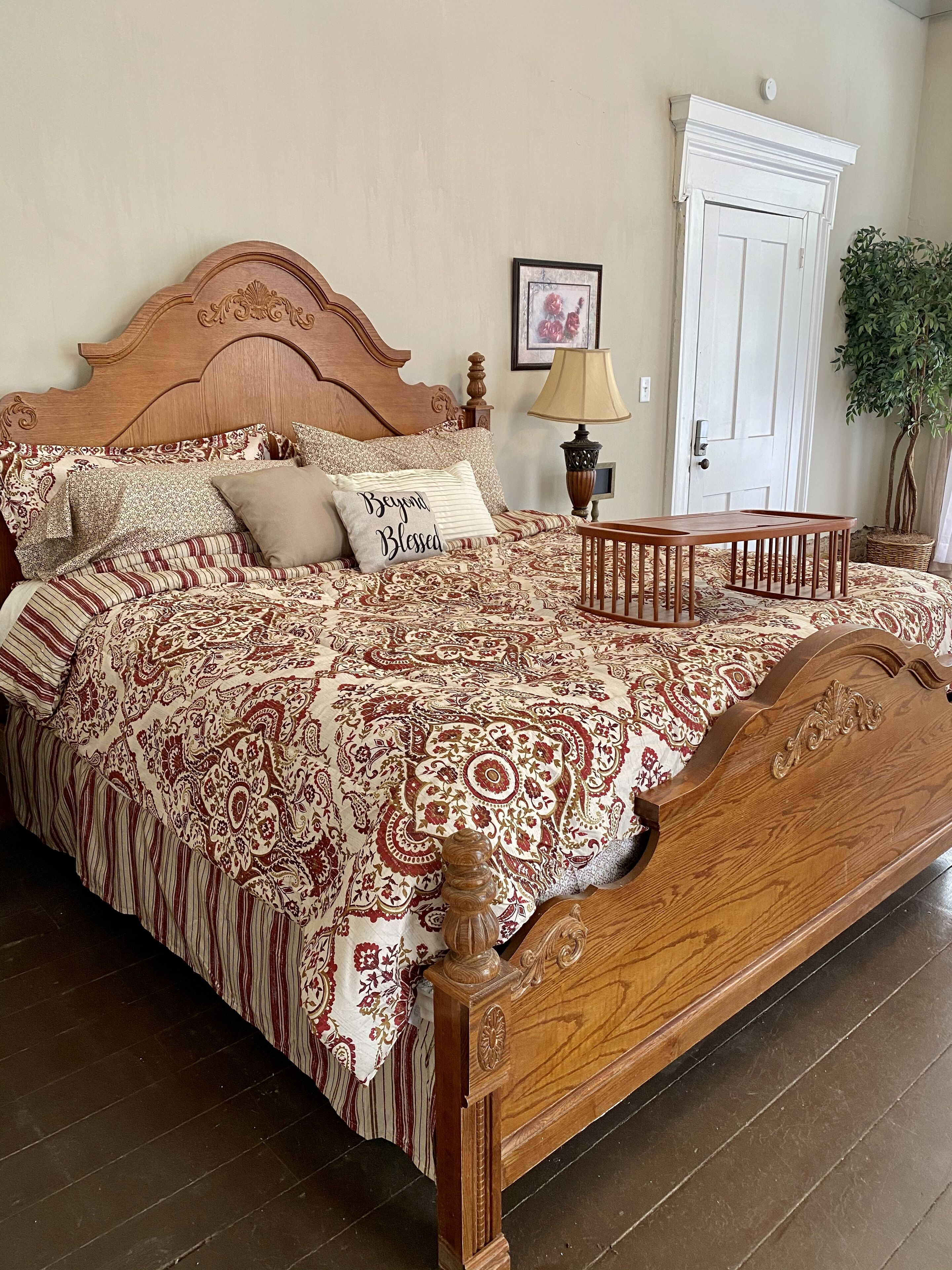 A wooden bed with a patterned bedspread, a bedside table with a lamp, a door, and a potted plant.
