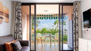 A modern living room with a sofa, a dining table with chairs, a TV, and a balcony with a view of palm trees and a clear sky.