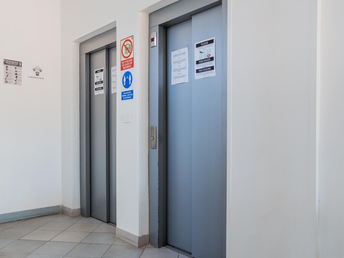 Un couloir avec deux ascenseurs, chacun étant muni d’un panneau indiquant qu’il est interdit de fumer et d’un indicateur de distance.