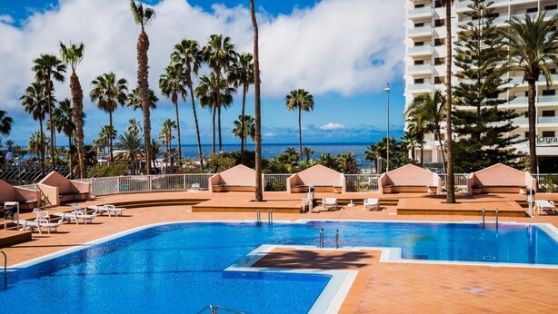 A swimming pool with lounge chairs and umbrellas, surrounded by palm trees and a view of the ocean.