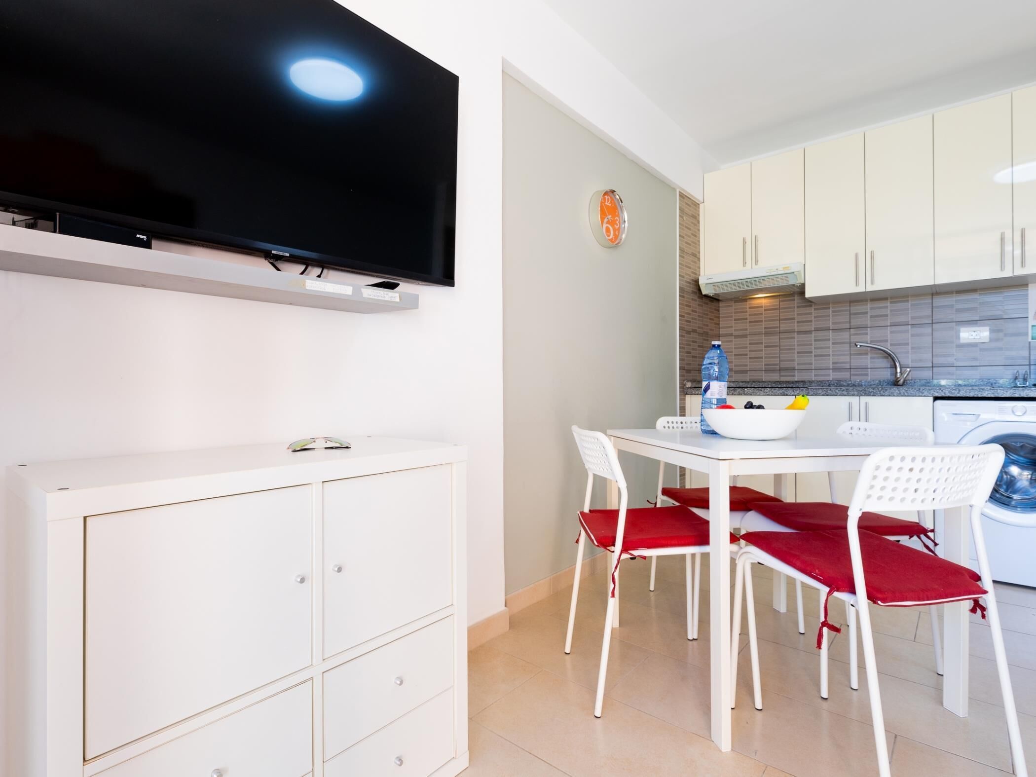 Una cuina moderna amb armaris blancs, un televisor de pantalla plana, una taula de dinar amb cadires vermelles i una rentadora.