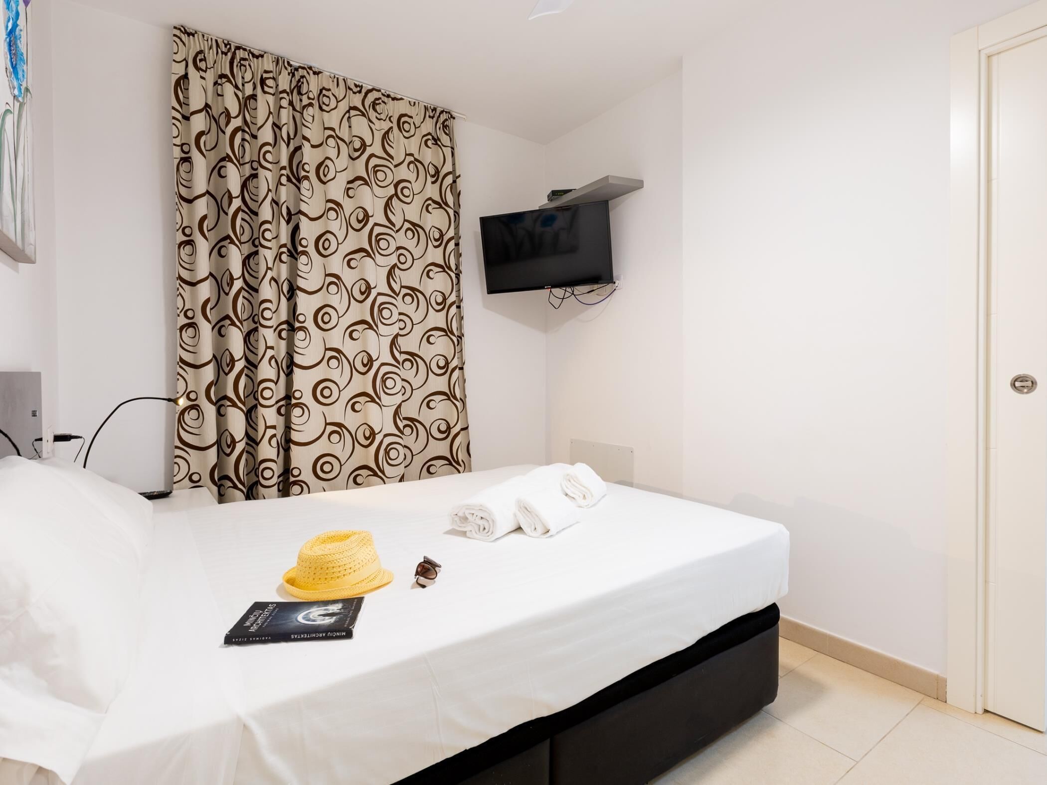 Quarto de hotel com cama, televisão fixada na parede, porta e cortina com padrão de redemoinho.