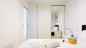 A hotel room with a bed, a bedside table with a lamp, a small desk with a chair, and a bathroom visible through an open door.