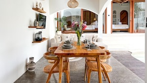 Un comedor con mesa de madera, sillas y un jarrón central con flores. Se ven estantes con objetos decorativos y una cama a través de un arco.