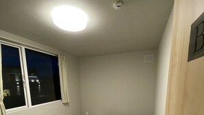 Una cama individual con ropa blanca en una habitación con ventana, una puerta con el letrero 'B' y una lámpara en el techo.