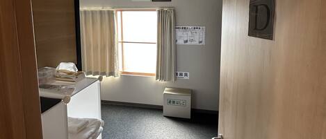 Una habitación de hotel con cama, escritorio, ventana con cortinas, lámpara de pared y puerta con cartel.