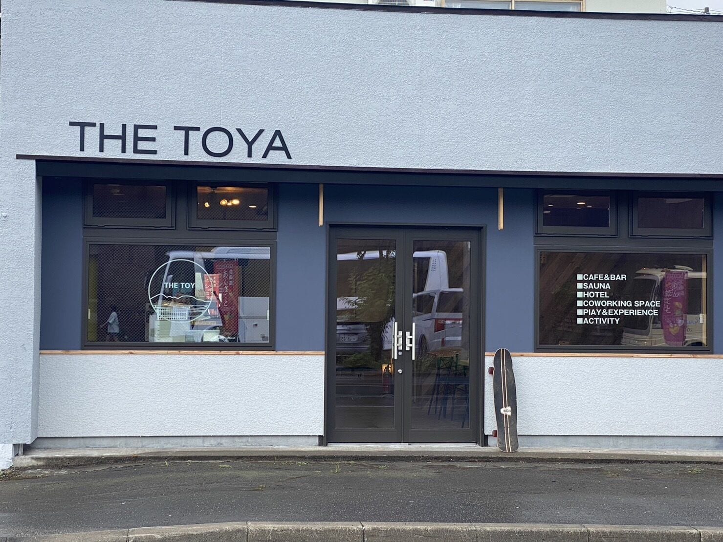 Een gebouw genaamd "THE TOYA" met een glazen deur en ramen die diverse voorzieningen tonen, zoals een café, bar, sauna, hotel, co-working ruimte, speel- en belevenisruimte, en activiteiten.