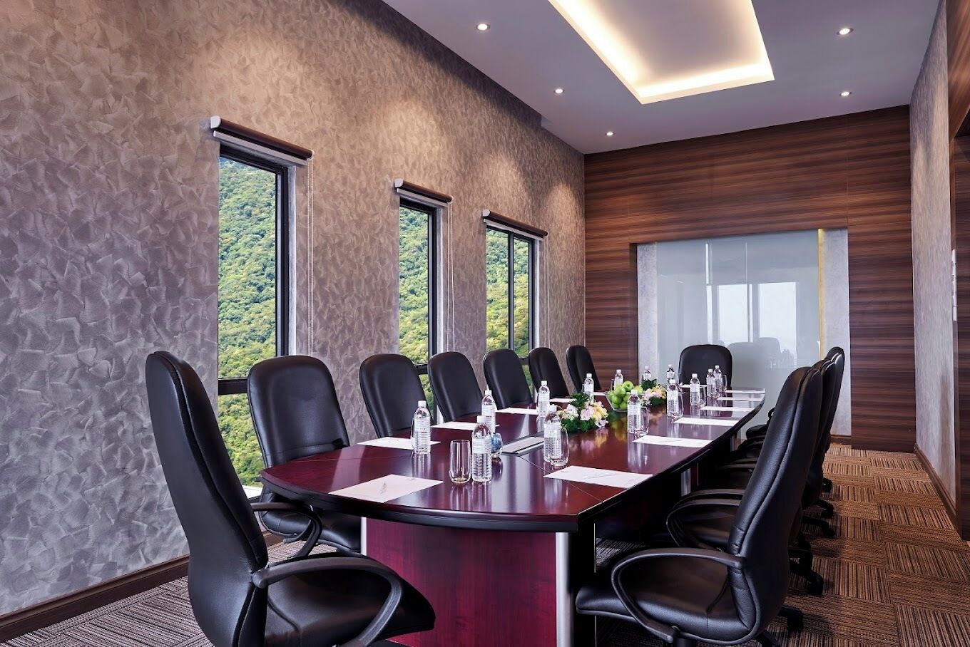 Ein Konferenzraum mit langem Tisch, schwarzen Stühlen und Blick auf Grün hinter großen Fenstern.