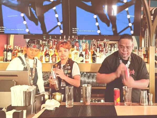 Un bar con un bartender preparando bebidas, una barra con diferentes botellas y vasos, y un fondo con estantes llenos de botellas.