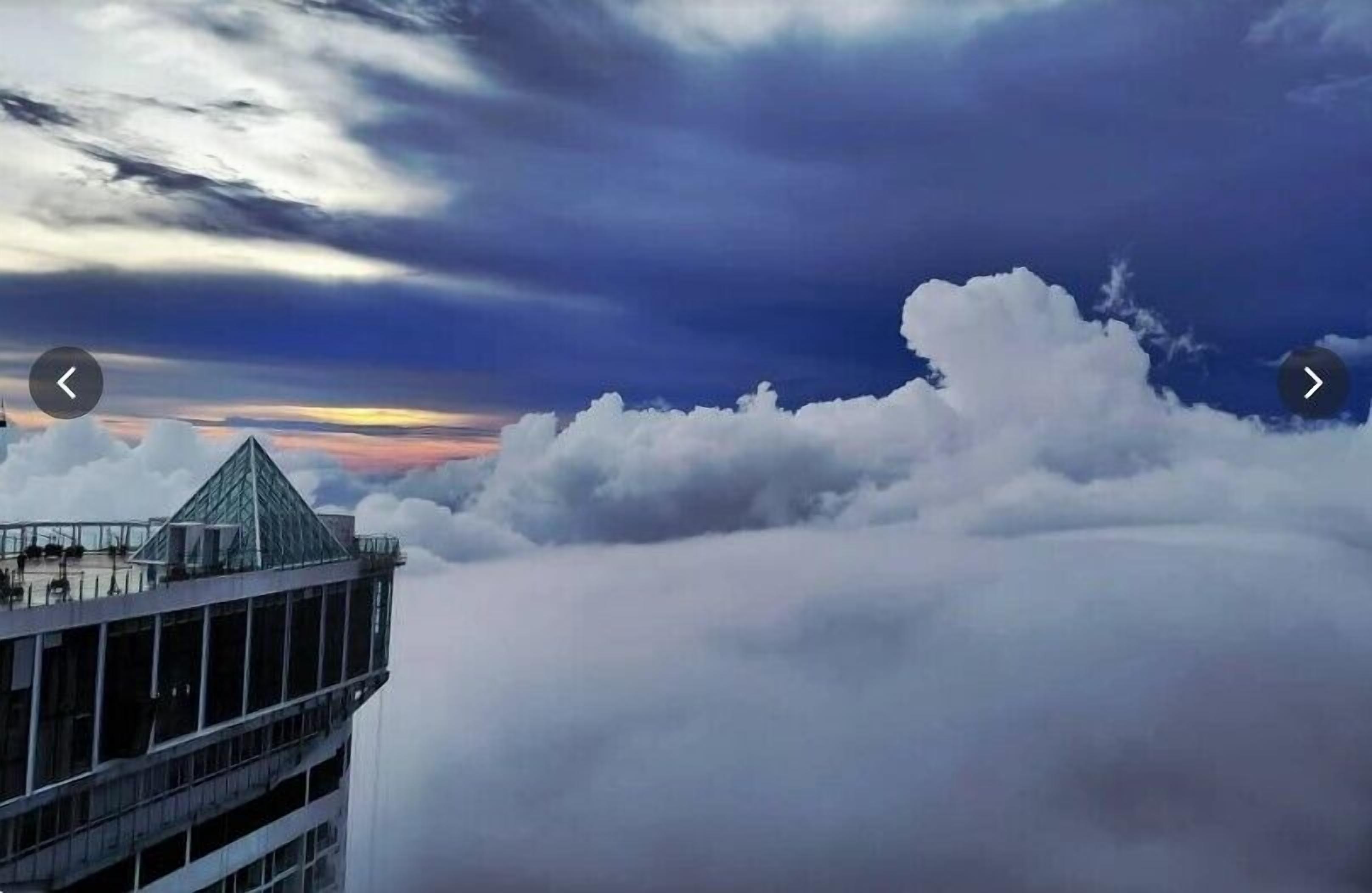 Um edifício alto com estrutura em formato de pirâmide de vidro e vista para as nuvens abaixo.