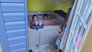 A hot tub with two people inside, a shelf with various items, and a window with blinds.