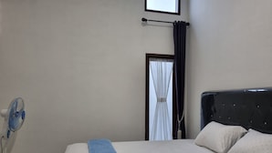 A bedroom with a bed, a standing fan, a window with curtains, and a door. - Wafaby Guest House (Payakumbuh)