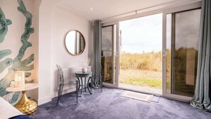 A modern room with a large sliding glass door leading to a grassy outdoor area, a round mirror on the wall, and a small round table with a lamp. - Magnificent Beach House - Seaview - Upside-down (Caister-on-Sea)