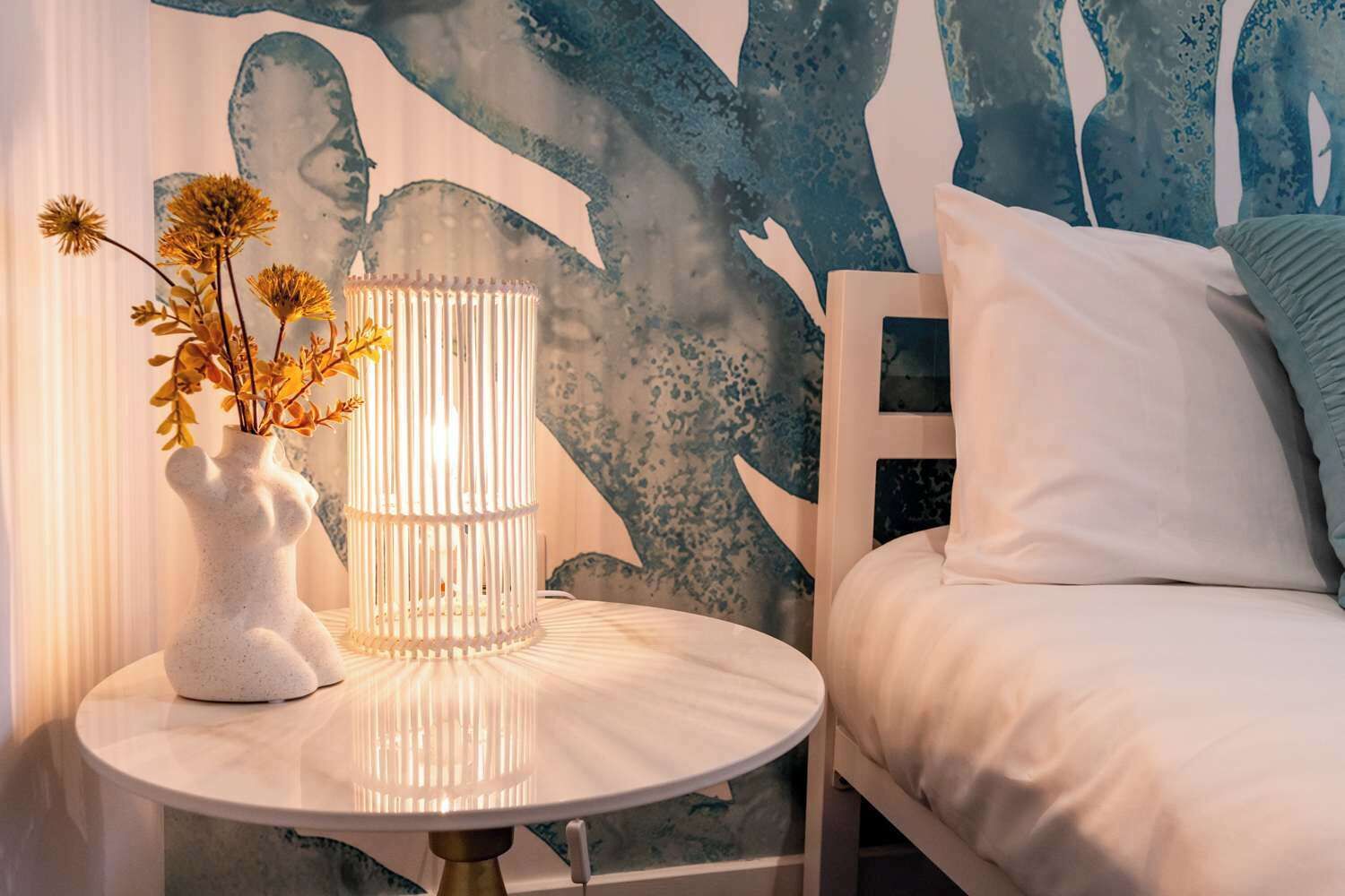 Une chambre à coucher avec un lit, une table ronde blanche ornée d’un vase et de fleurs, et une fresque murale.