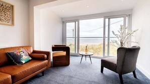 A modern living room with a leather sofa, a brown armchair, and a round table with a bottle and glasses. Large windows offer a view of the beach. - Magnificent Beach House - Seaview - Upside-down (Caister-on-Sea)