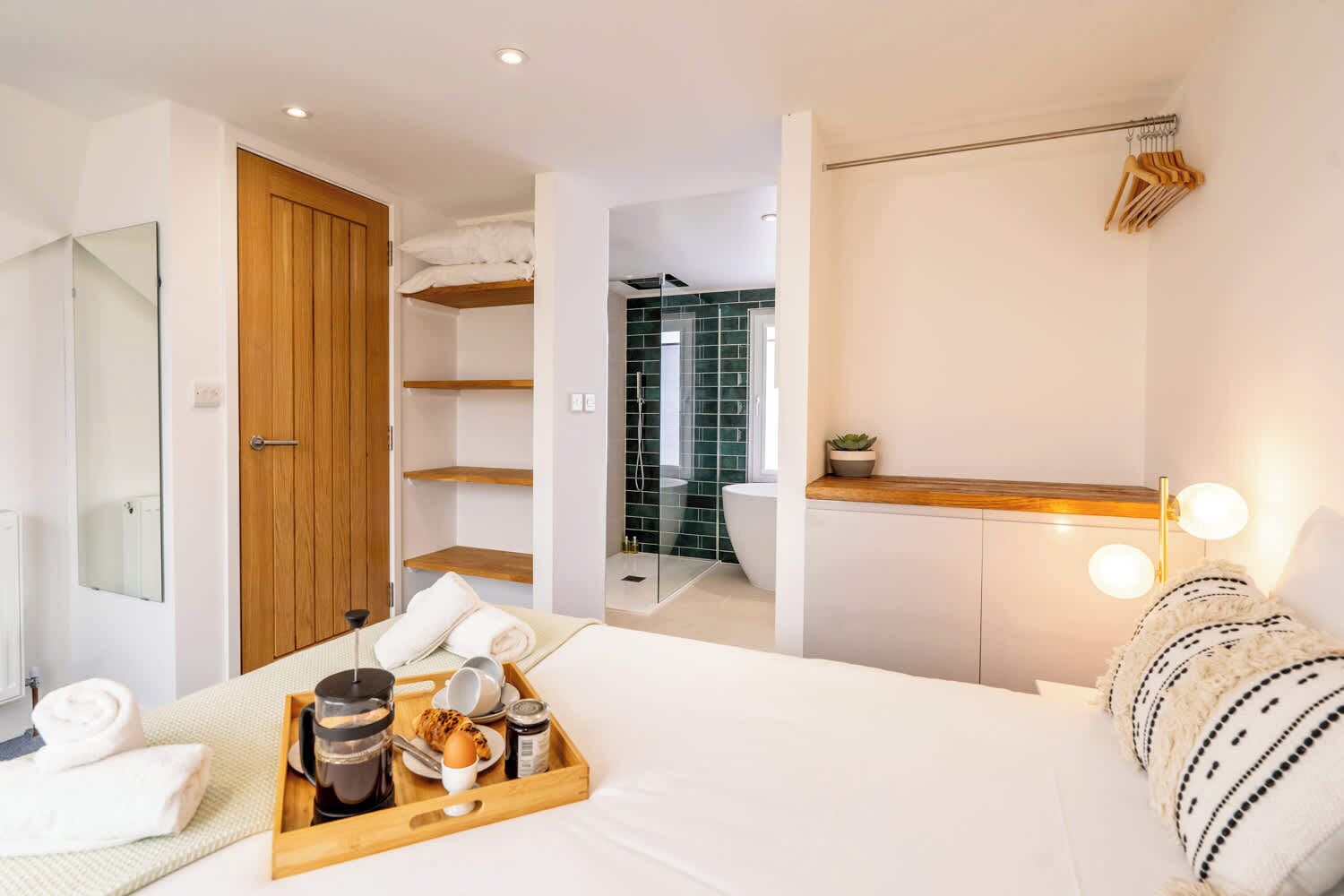 A modern hotel room with a bed, a wooden wardrobe, a bathroom with a glass shower, and a breakfast tray on the bed.