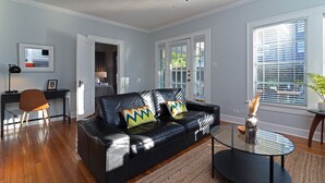 A modern living room with a black leather sofa, a glass coffee table, and a ceiling fan. - 3BR The Poseidon! Aquatic Dream Pool in Montrose! (Houston)