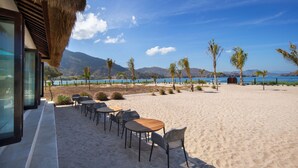 A beachside restaurant with outdoor seating and a view of the ocean and mountains. - Mocean Beach Resort (Sumbawa Barat)