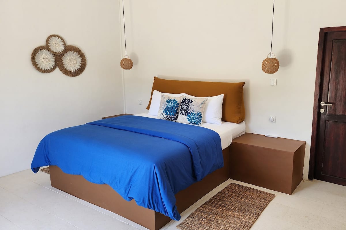 A bedroom with a bed, a wooden headboard, a brown bedside table, a blue bedspread, and a woven rug.