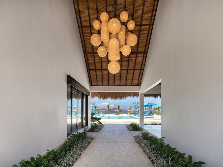 Un couloir mène à un espace piscine doté d’un toit de chaume et de luminaires sphériques suspendus.