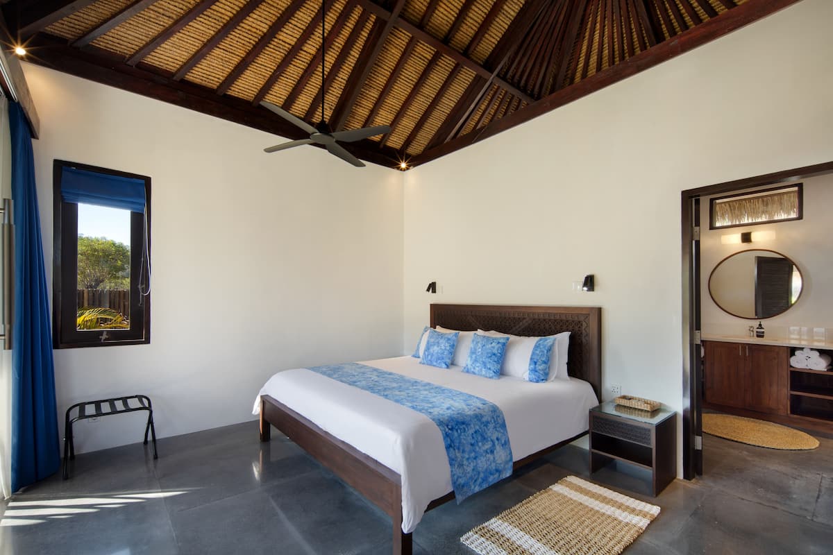A bedroom with a bed, a ceiling fan, a nightstand, a bench, a bathroom with a mirror, and a door.