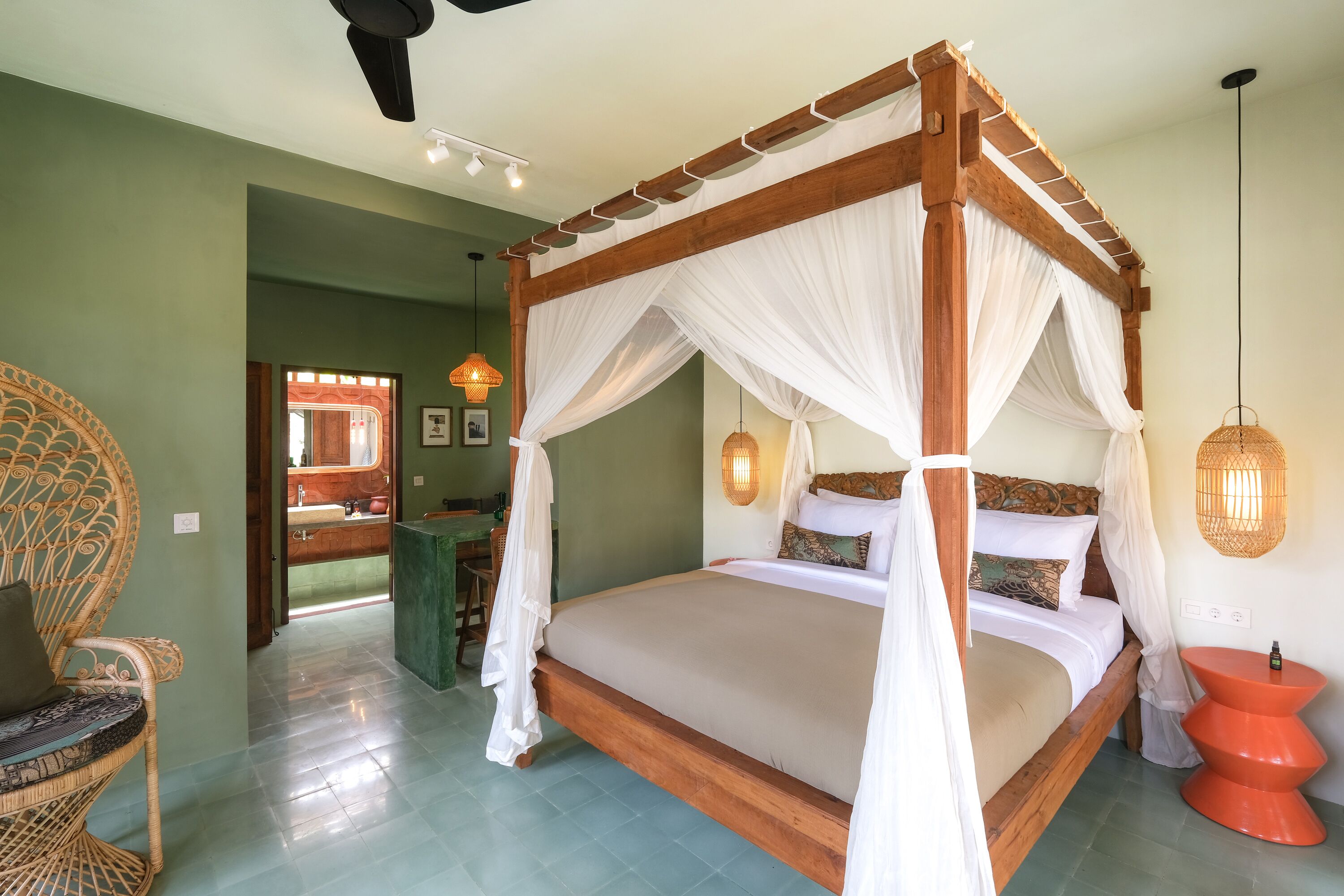 A four-poster canopy bed with a wooden frame and white drapes, a wicker chair, and a red round table with a candle.