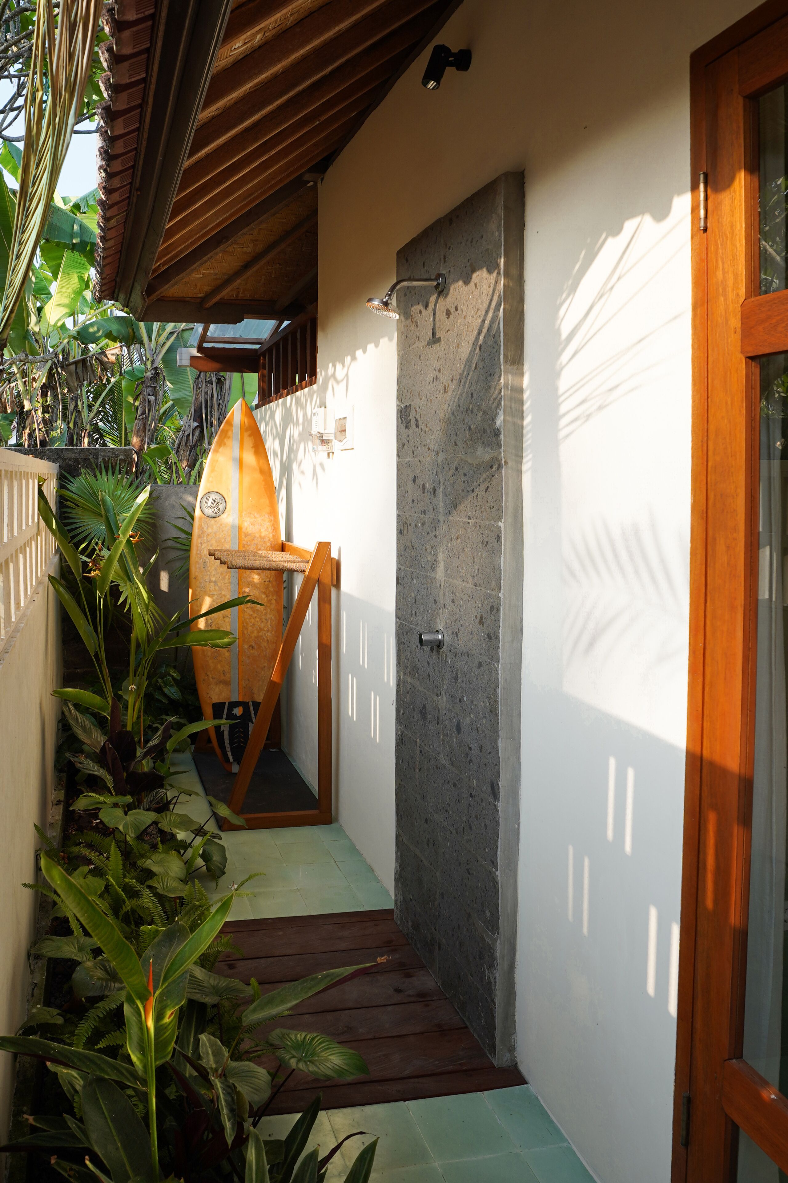 A narrow hallway with a surfboard leaning against the wall, a wooden door, and a concrete wall.