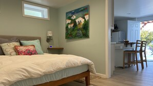 Ein Schlafzimmer mit einem Bett, Kissen, einem Nachttisch, einem Gemälde und Blick in einen Küchenbereich.