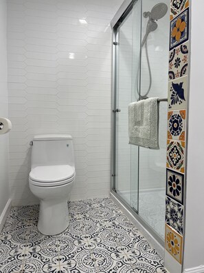 Casa de banho com sanita branca, cabine de duche com portas de vidro e chuveiro manual, e chão com padrão.