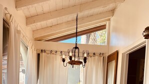 Une pièce chaleureuse avec un plafond en bois, un lustre, une table, une chaise et une vue sur l’extérieur à travers une fenêtre ornée de rideaux.