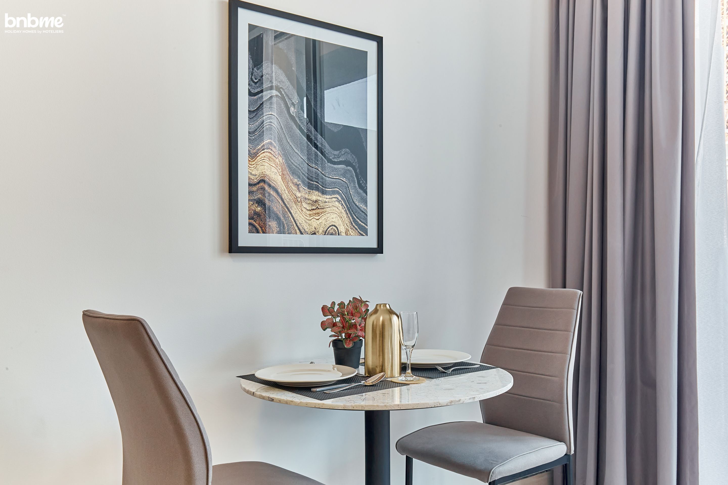 A dining area with a round table, two chairs, a framed artwork, a potted plant, and a vase.