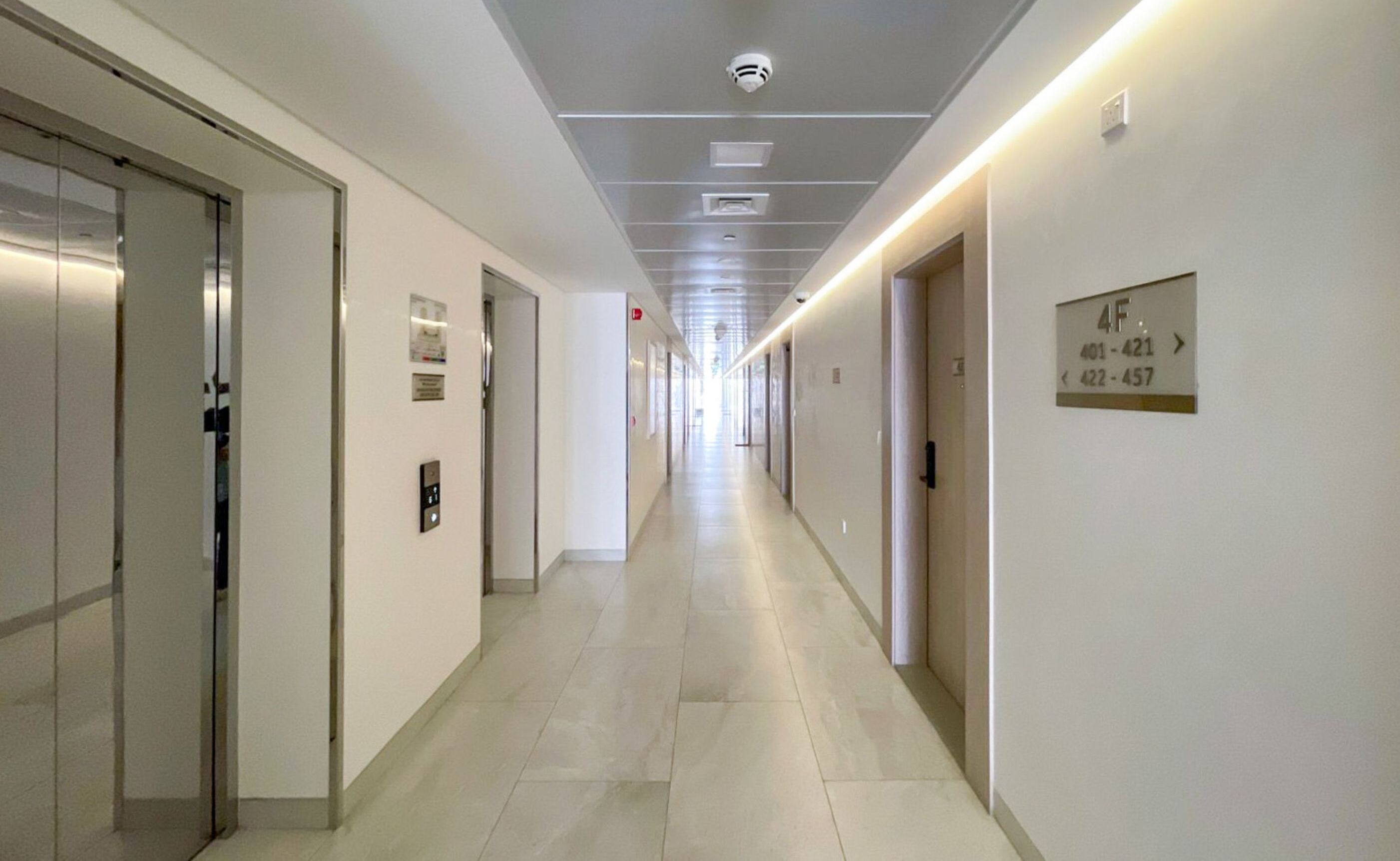 Un long couloir bien éclairé, avec des portes de chaque côté, un plafond doté d’un éclairage encastré et un panneau indiquant les numéros des pièces.
