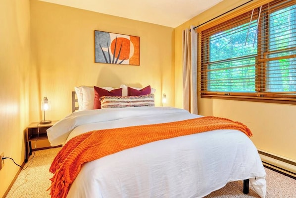 A bedroom with a bed, a nightstand, a lamp, a window with blinds, and a wall art piece.