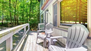 A wooden deck with Adirondack chairs, a wooden railing, and a window with blinds overlooking a forested area. - Tranquil Grove | Hot Tub | Game Room | Fire Pit (Lake Ariel)