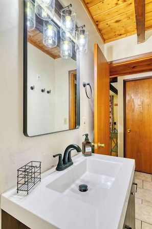 Une salle de bain avec un grand miroir, un lavabo blanc et un robinet noir. Il y a un luminaire au-dessus du miroir et une porte sur la droite.