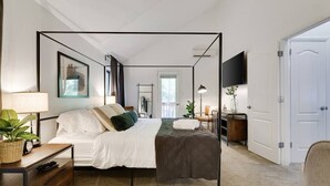 Une chambre moderne avec un lit à baldaquin, une table de chevet avec une lampe, un téléviseur fixé au mur et une porte donnant sur une autre pièce.